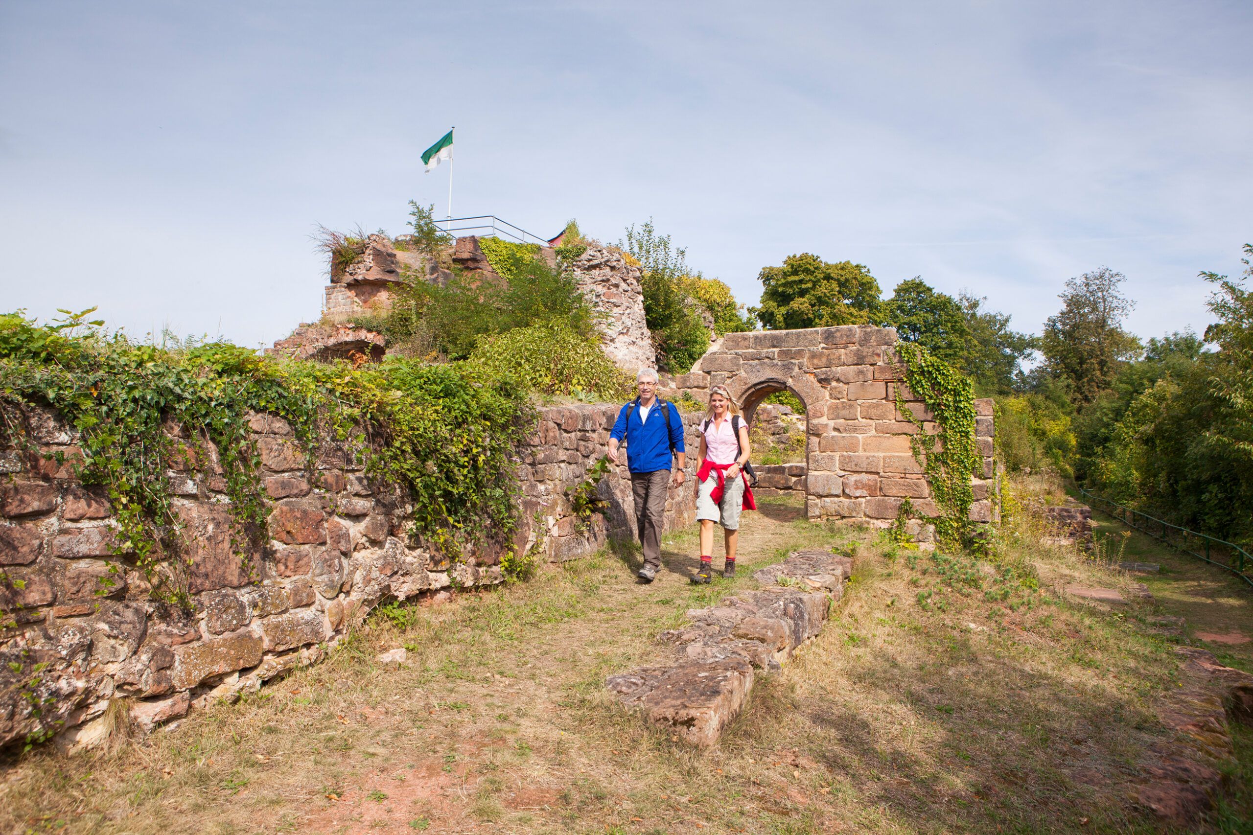 Zwei Wandernde auf dem Pfad zwischen den Sandsteinmauern der Ruine Hohenburg, dahinter ein Torbogen und eine Fahne