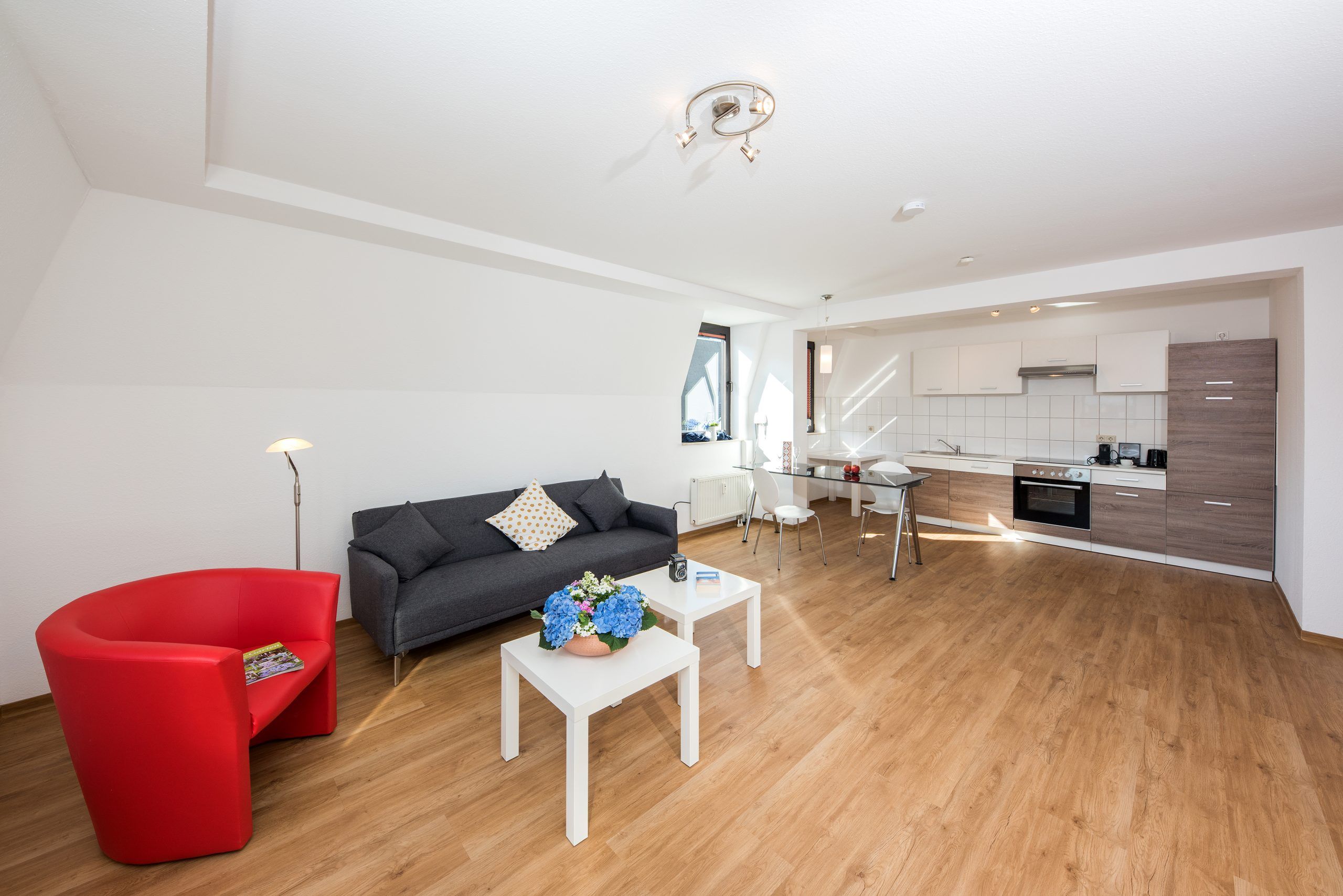 Heller Wohnbereich eines Apartments der City Residenz mit rotem Sessel, grauem Sofa und offener Küchenzeile