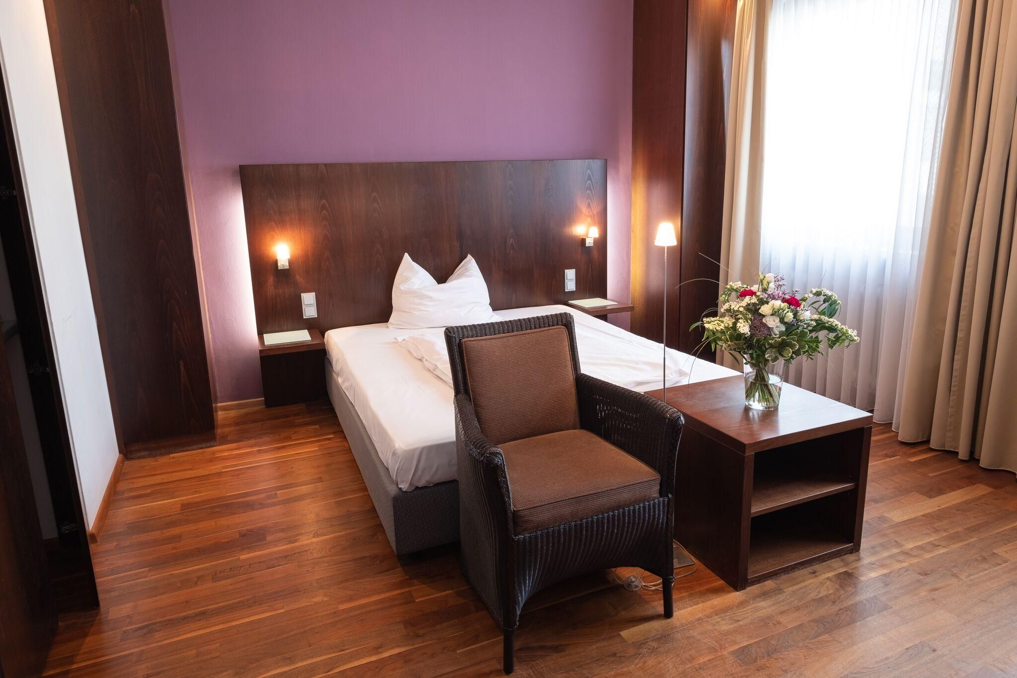 Studio der City Residenz mit Doppelbett vor fliederfarbener Wand, dunklem Holzkopfteil, Korbsessel und Parkett