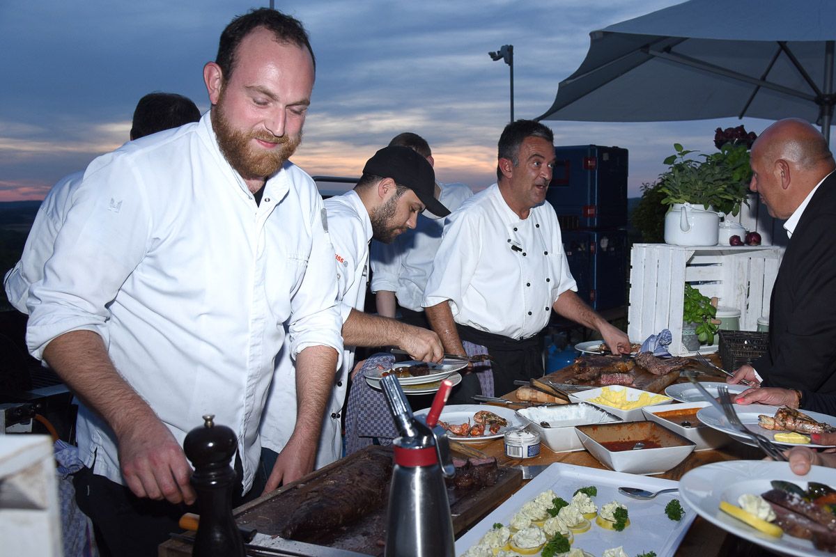 Köche in weißen Jacken tranchieren Fleisch am Buffet im Freien, dahinter der Abendhimmel über dem Tal