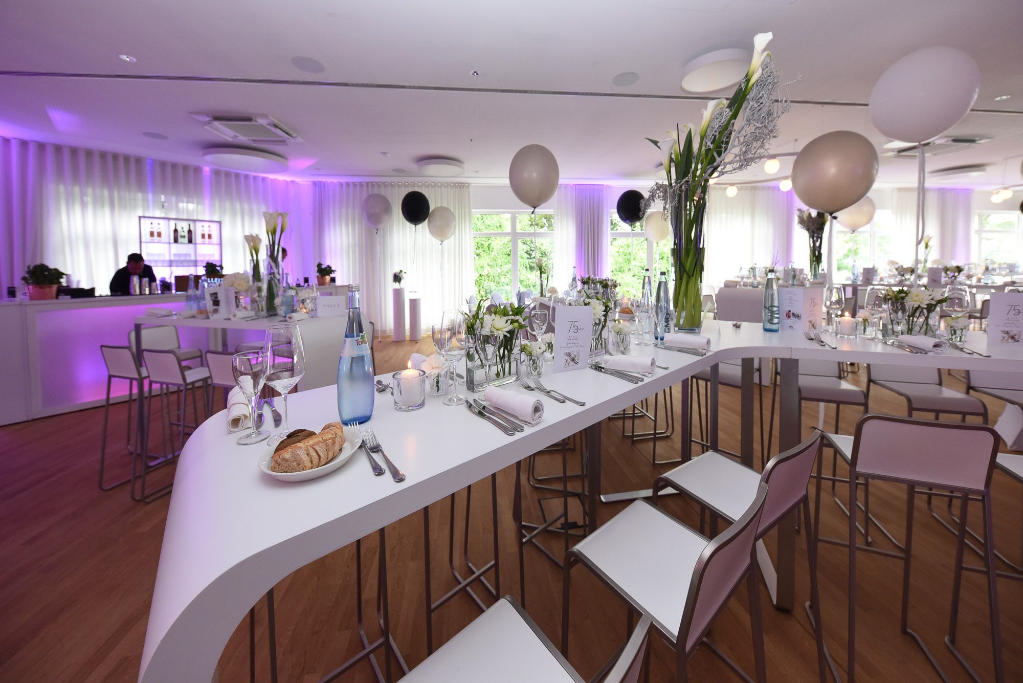 Geschwungene weiße Hochtische mit Barhockern, Ballons und violetter Beleuchtung im Saal, hinten die Bar