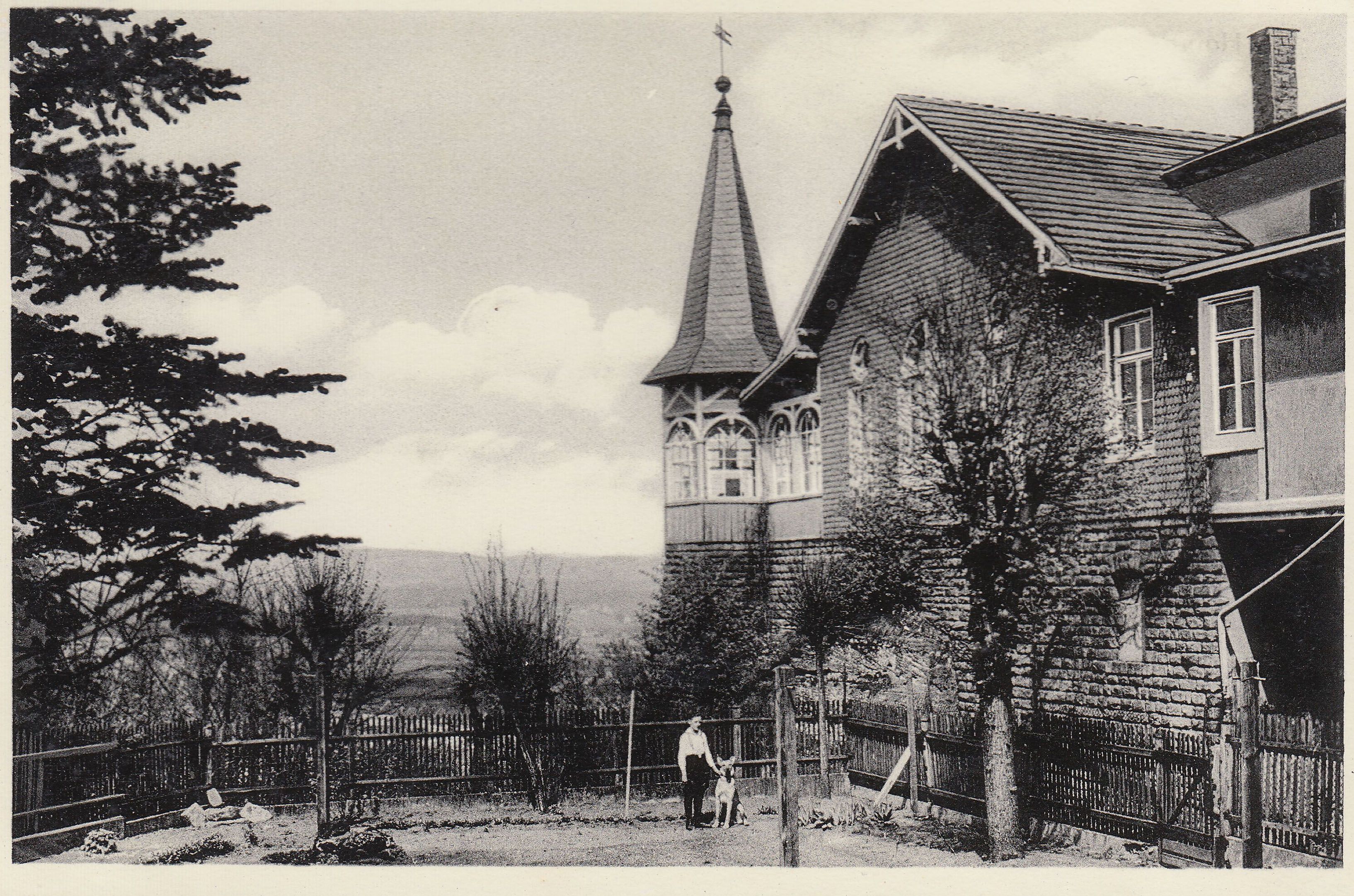 Historical black-and-white photo of a house with a pointed turret, a man with a dog by the wooden fence