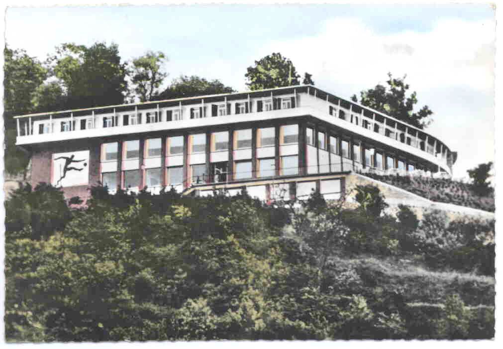 Tinted postcard of a long flat-roofed building on a wooded slope, with a relief of a footballer on the façade