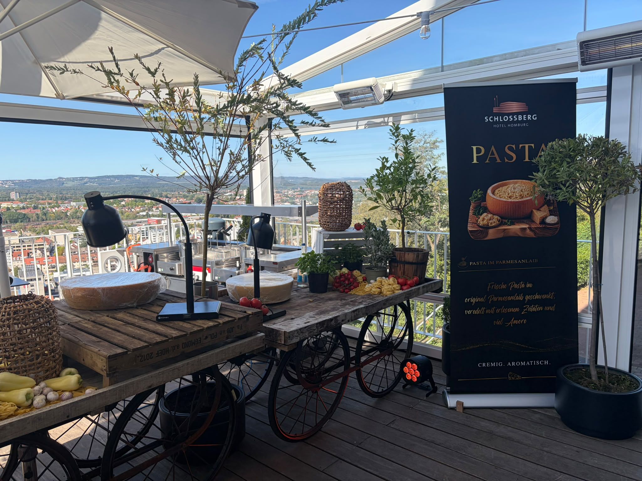 Pasta-Station mit Parmesanlaib auf einem Holzwagen auf der Terrasse, daneben ein Rollup „Pasta im Parmesanlaib“, im Hintergrund Homburg