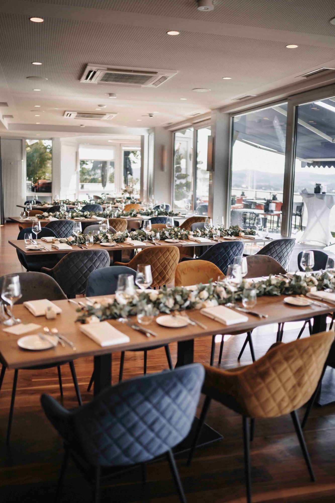 Restaurant festlich für eine Hochzeit eingedeckt: lange Tafeln mit Eukalyptus-Girlanden, Samtstühle und eine große Fensterfront