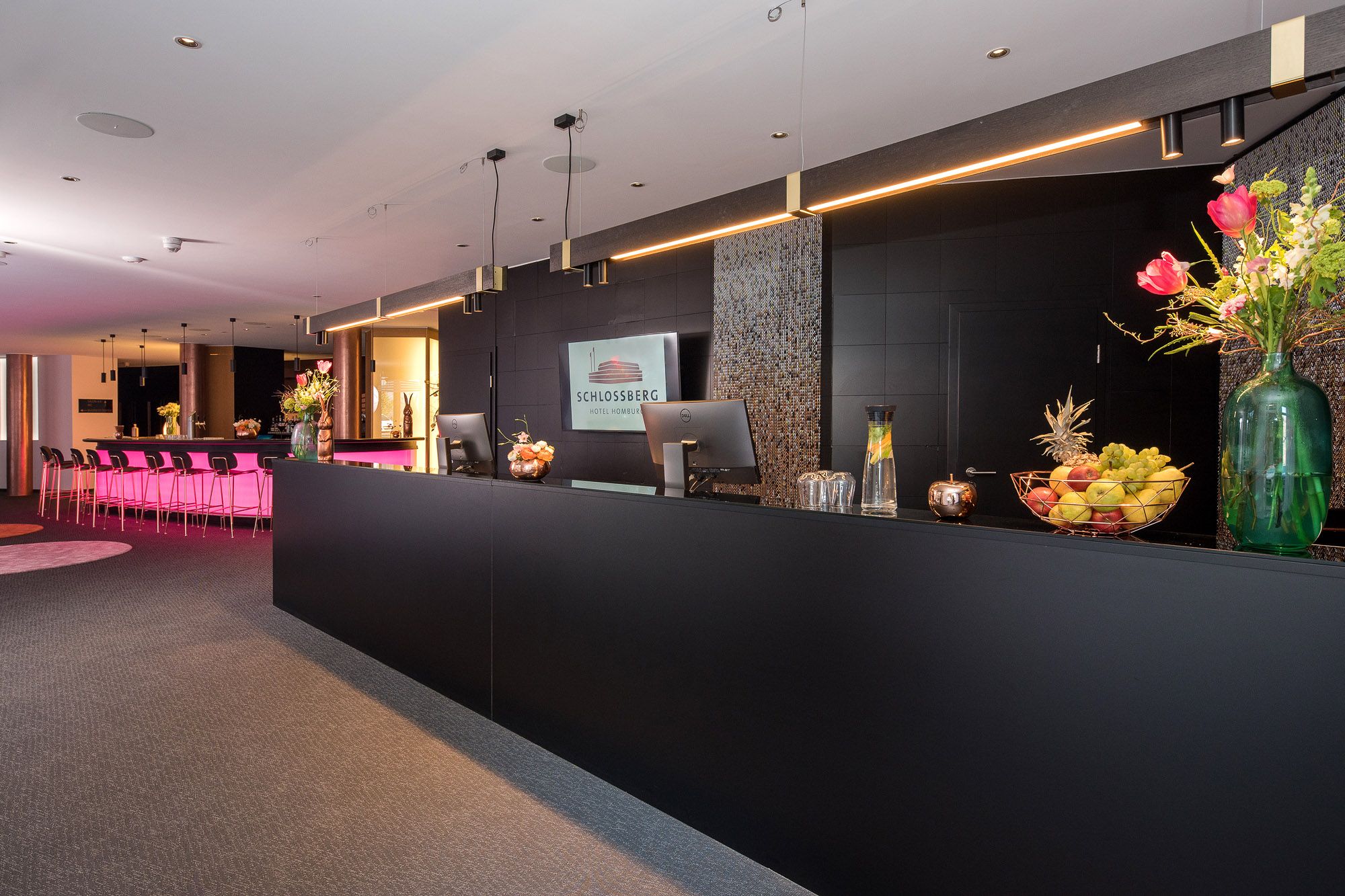 Schwarzer Empfangstresen mit Obstschale und Blumen in der Lobby, im Hintergrund die pink beleuchtete Bar