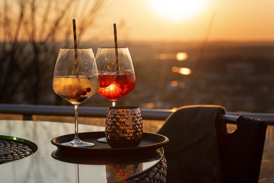 Zwei Aperitifs mit Strohhalm auf einem Tablett an der Terrassenbrüstung, dahinter die tief stehende Sonne über dem Tal