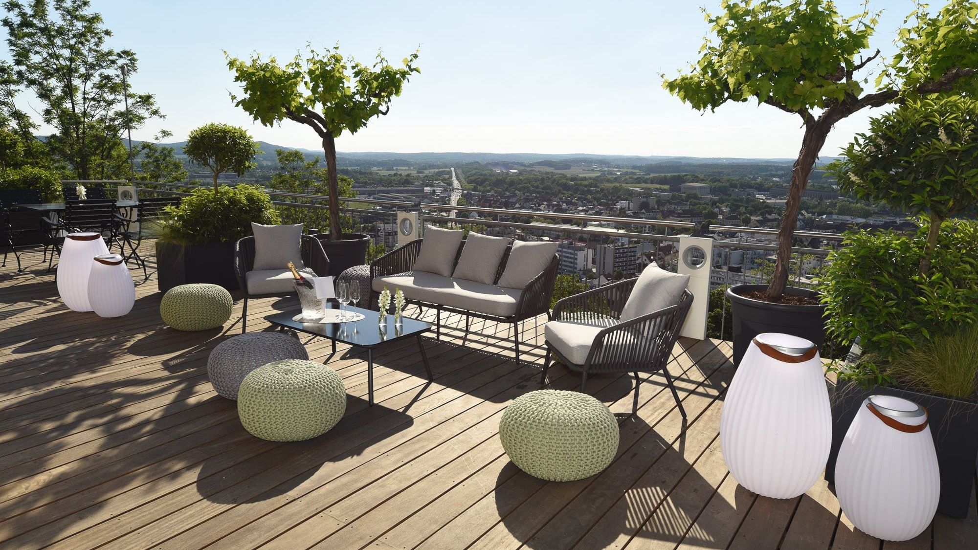 Lounge-Möbel, Strickhocker und Leuchtvasen auf der Holzterrasse, dahinter der Blick über die Dächer von Homburg