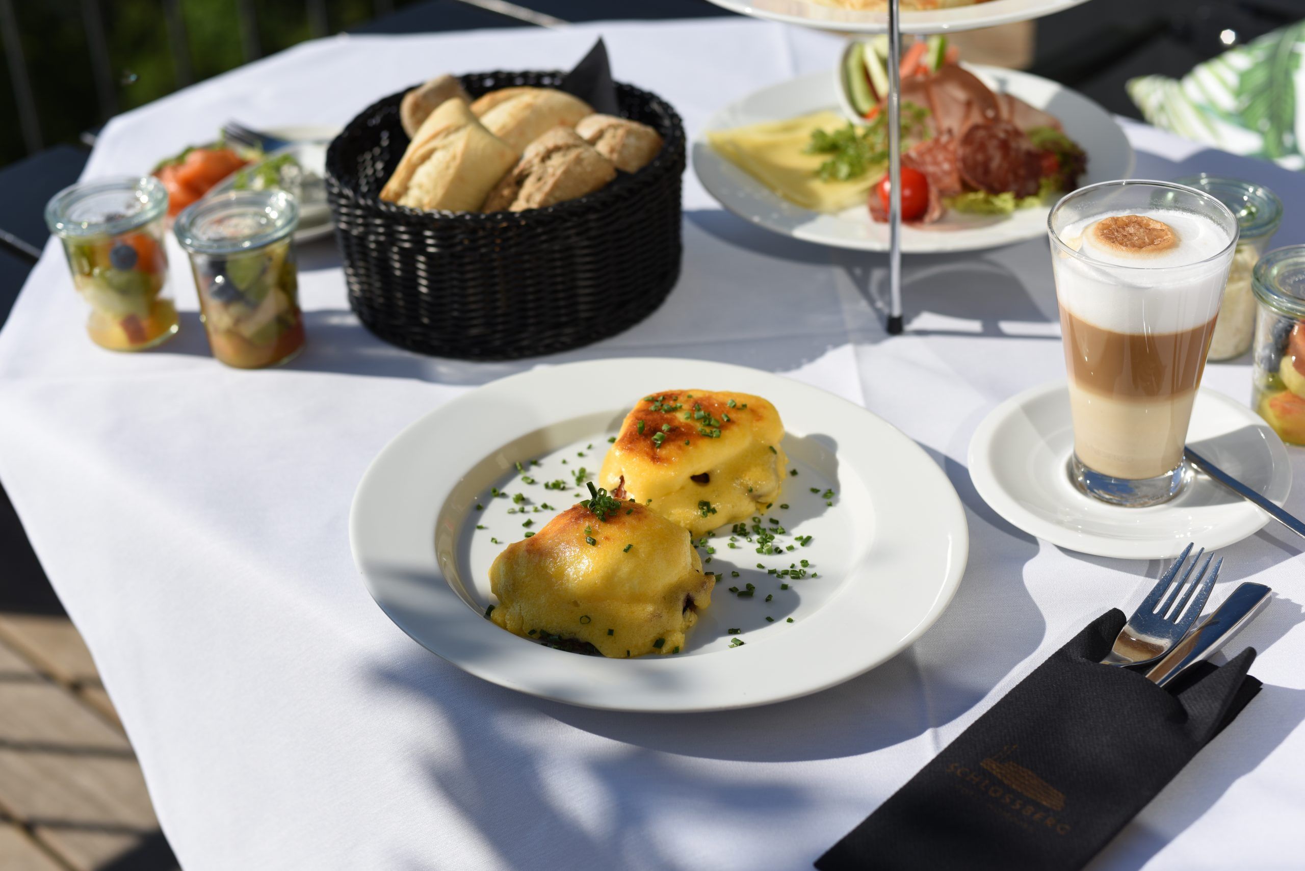Frühstückstisch in der Sonne mit überbackenem Gericht, Brotkorb, Latte macchiato, Obstsalat im Glas und Aufschnitt