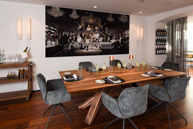 Gedeckte Holztafel mit Kupferlampen und grauen Samtstühlen vor einem großen Schwarz-Weiß-Wandbild, rechts ein Weinregal