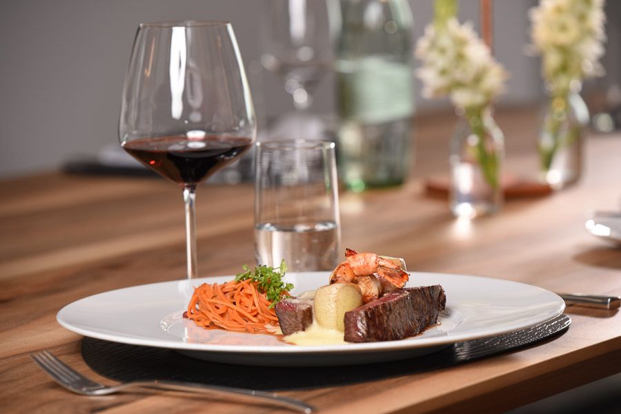 Tranchiertes Rindersteak mit Garnelen, Möhrenstreifen und Kartoffel auf weißem Teller, dahinter ein Glas Rotwein