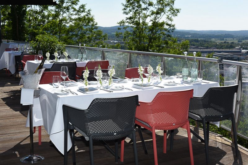 Weiß eingedeckte Tische mit roten und schwarzen Stühlen auf der Terrasse, dahinter Bäume und bewaldete Hügel