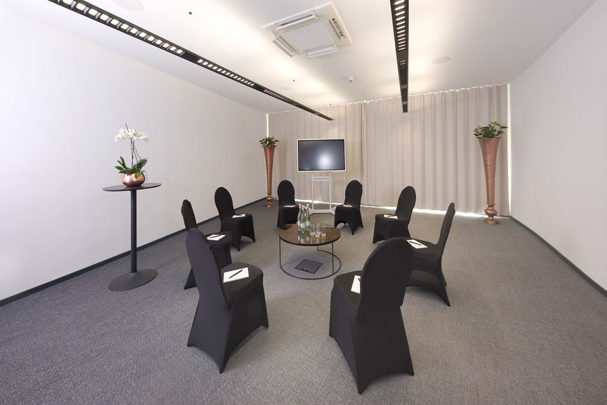 Meeting room with black-covered chairs in a circle around a glass table, a screen and light curtain behind