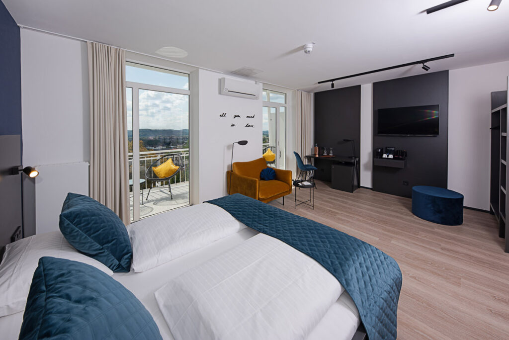 Bellevue Deluxe Zimmer mit sicht ins Zimmer auf Wand mit TV und Bett. Balkon direkt auf der linken Zimmerseite mit Bestuhlung