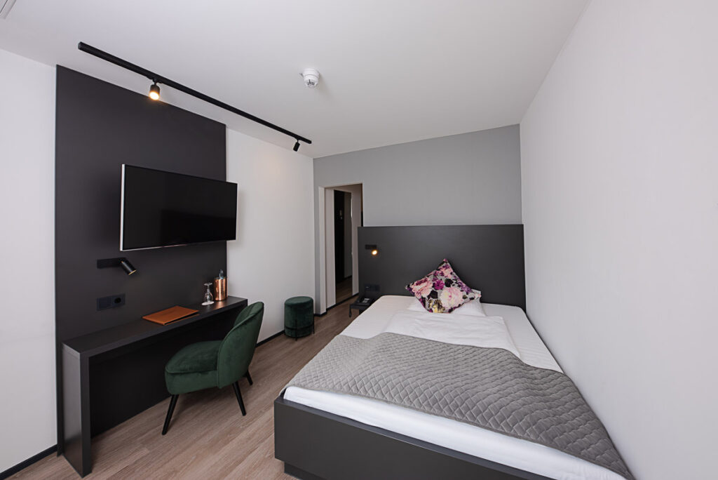 Business Einzelzimmer von ca. 16 qm mit Queensize Bett mit Blick auf das Bett und auf der linken Seite ist ein Schreibtisch mit TV und Sessel