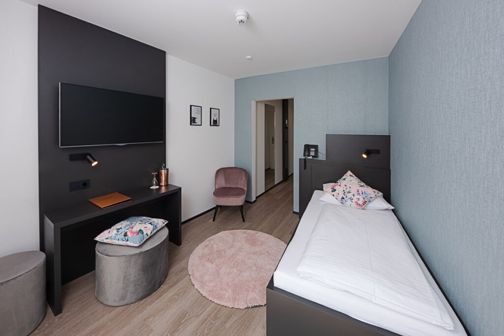 Comfort Einzelzimmer von ca. 14qm mit Queensize Bett mit Blick auf das Bett und auf der linken Seite ist ein Schreibtisch mit TV und Sessel