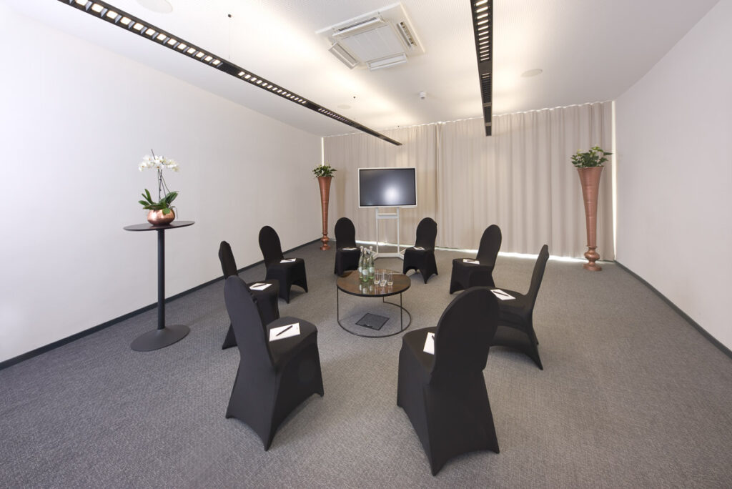 Konferenzraum mit Bestuhlung und Stehtischen. Die Stühle sind mit schwarzen Hussen bezogen und sind zu einem Kreis gestellt