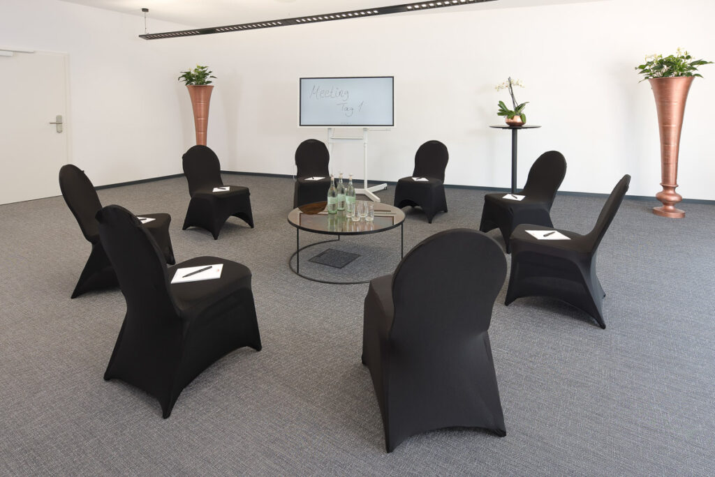 Konferenzraum mit Bestuhlung und Stehtischen. Die Stühle sind mit schwarzen Hussen bezogen und sind zu einem Kreis gestellt im Hintergrund steht ein Whiteboard