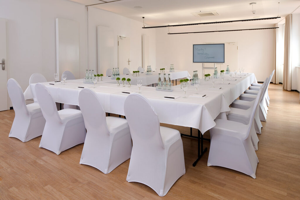 Konferenzraum mit weißer Bestuhlung und dekorierten Tischen. Die Tische sind als U aufgestellt auf den Tischen stehen kleine Wasserflaschen und Vasen mit Blumen