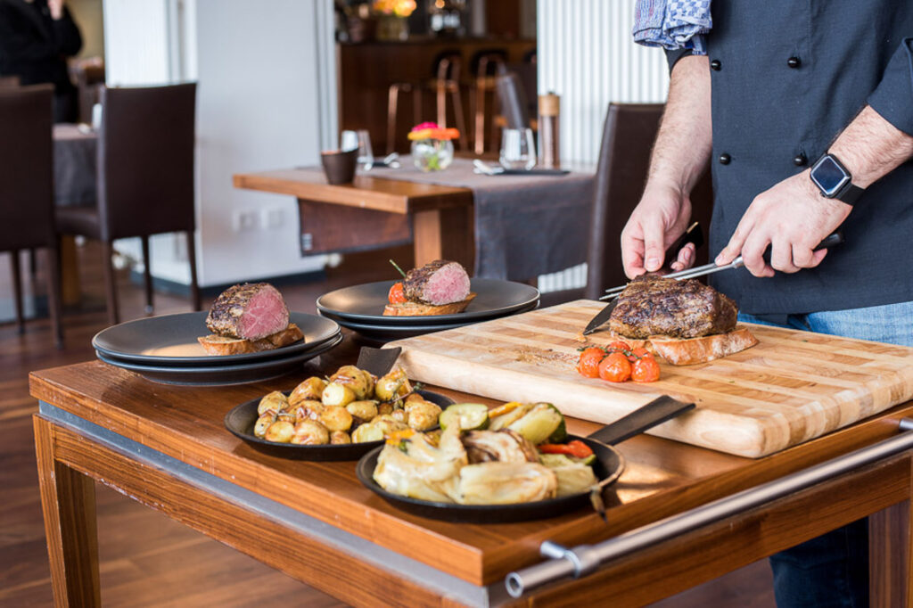 Koch schneidet ein Stück Fleisch frisch gebraten auf einem Holzbrett und richtet es auf einer gerösteten Scheibe Brot an mit Rosmarinkartoffeln und gegrilltem Sommer Gemüse